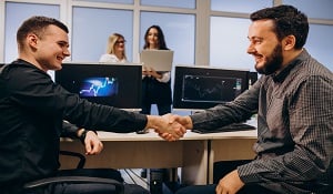 Two teammates in front of their worskstations and  shaking their hands while two other gilrs standing behind the workstations and looking into their laptop screen.