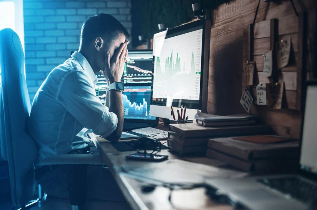 A man sitting in front of a desktop with his face covered with his boh hands and looks stressed - Cyber security is failing